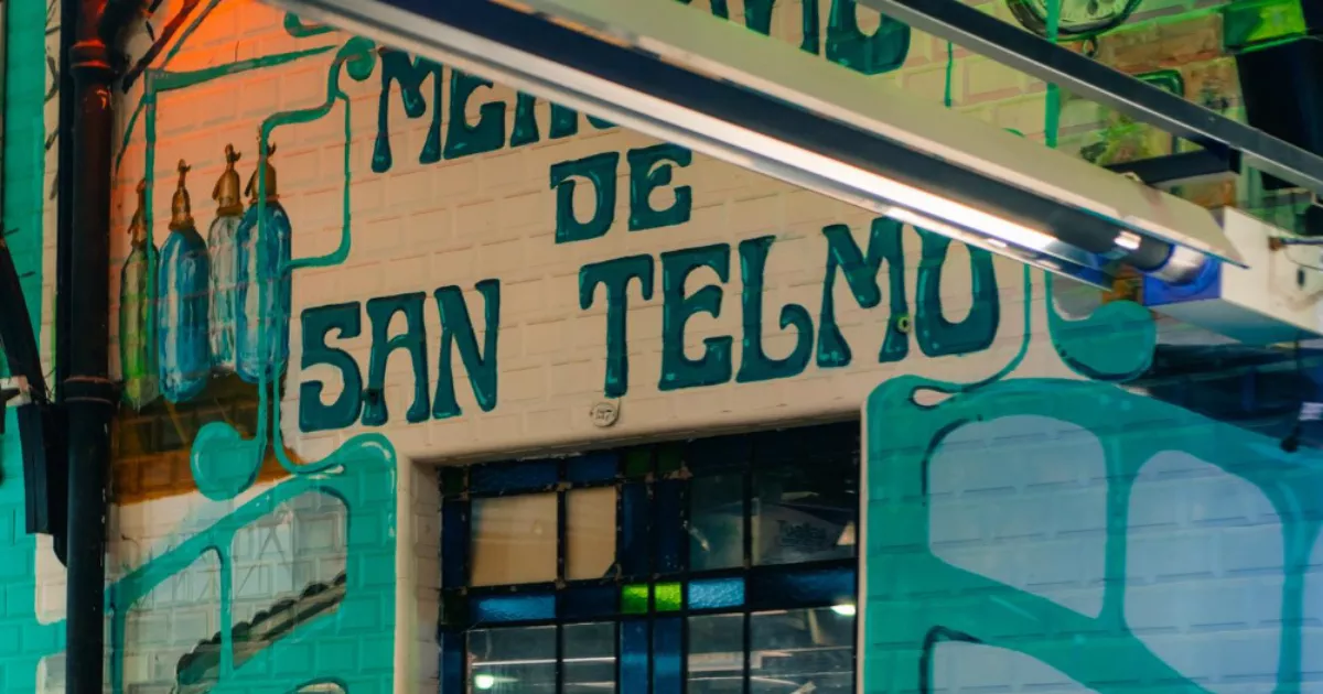 Fachada de bar iluminada à noite com letreiro pintado à mão em letras verdes sobre parede de tijolos brancos e grafite colorido na entrada do estabelecimento