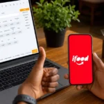 Mão segurando celular com o aplicativo iFood aberto ao lado de notebook exibindo sistema PDV com cardápio e preços, sobre mesa de restaurante