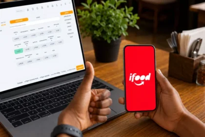 Mão segurando celular com o aplicativo iFood aberto ao lado de notebook exibindo sistema PDV com cardápio e preços, sobre mesa de restaurante