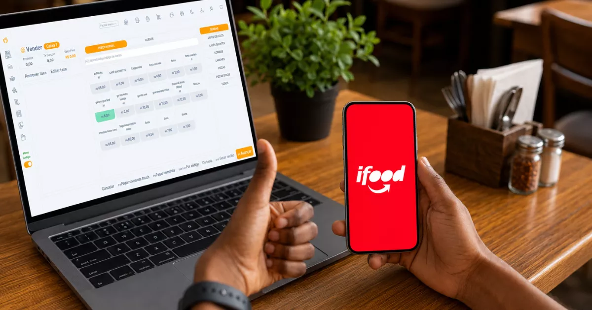 Mão segurando celular com o aplicativo iFood aberto ao lado de notebook exibindo sistema PDV com cardápio e preços, sobre mesa de restaurante