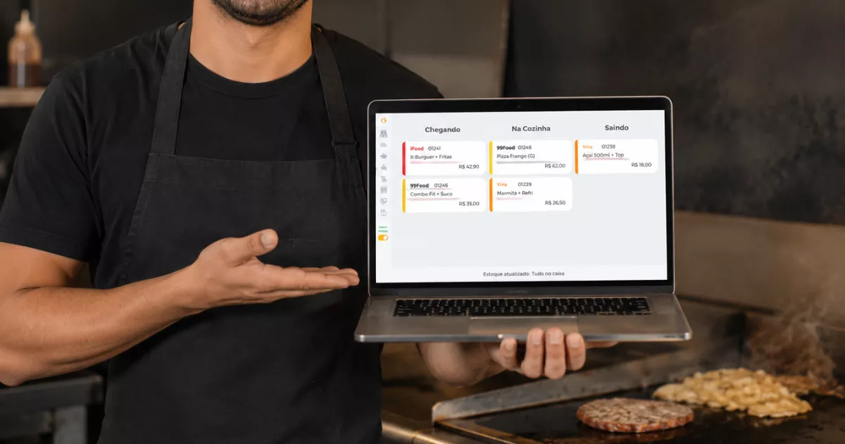 Cozinheiro de avental preto apresentando laptop com a tela do KDS do ControleNaMão exibindo pedidos do iFood, 99Food e Vina organizados por status, ao lado da chapa com hambúrgueres