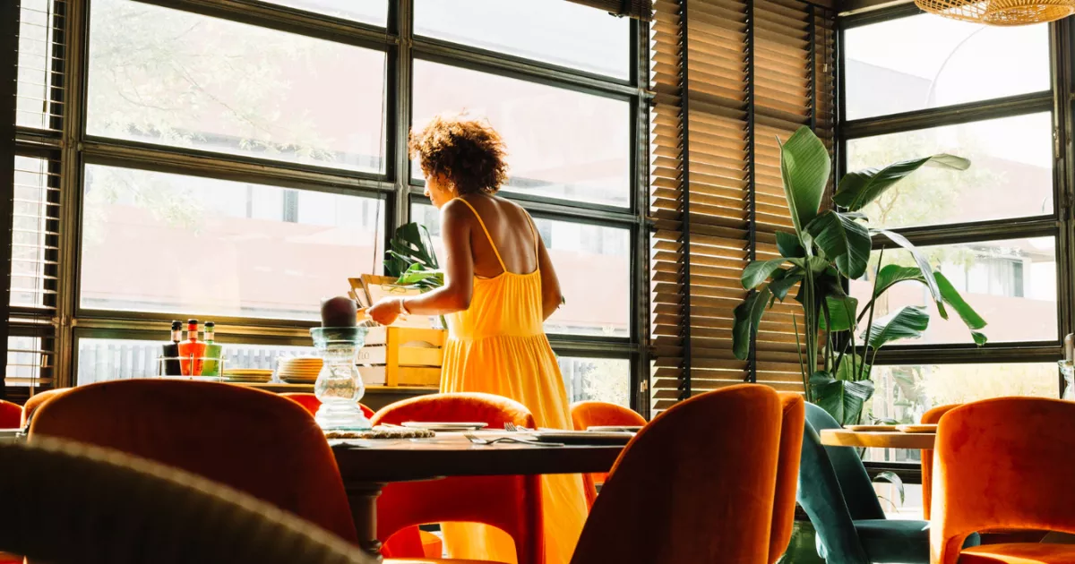 Salão de restaurante com luz natural quente da tarde, cadeiras de veludo laranja, plantas tropicais e funcionária de vestido amarelo organizando mesa antes da abertura