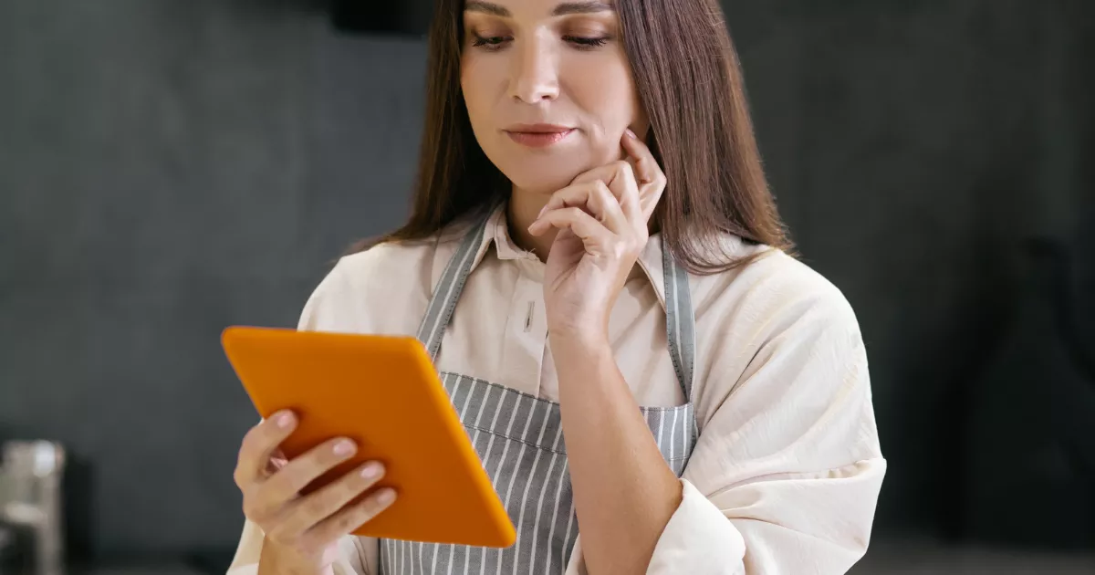 Gestora de avental listrado consulta tablet com a mão no queixo, analisando dados na cozinha do restaurante para controlar custos de produção