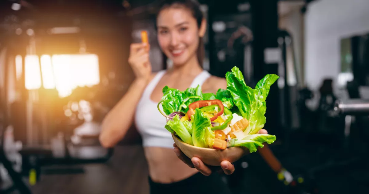 Mulher sorrindo em roupa de academia segurando tigela com salada colorida dentro de ginásio, ilustrando parceria entre negócio de alimentação e academia para atrair clientes