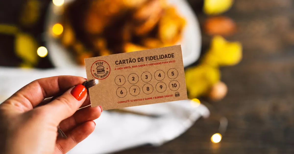 Mão feminina segurando cartão de fidelidade kraft com dez campos numerados e texto "complete 10 visitas e ganhe 1 burger grátis", ao fundo prato com comida e luzes desfocadas