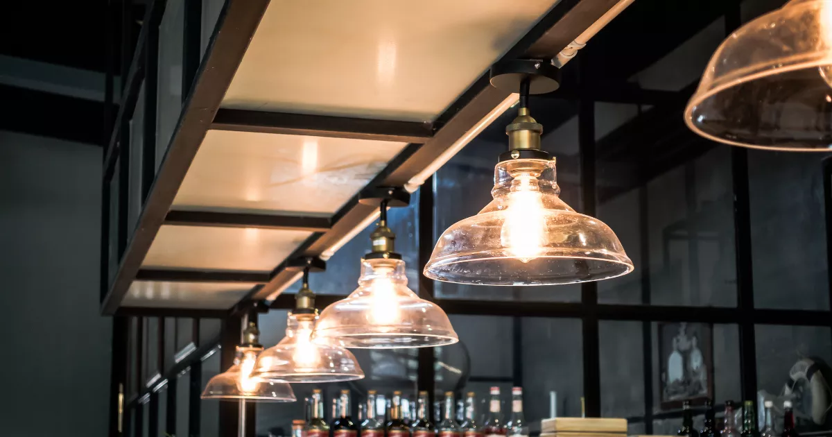 Três pendentes de vidro transparente acesos sobre o balcão de um bar com garrafas ao fundo, iluminação que impacta o custo de energia do restaurante