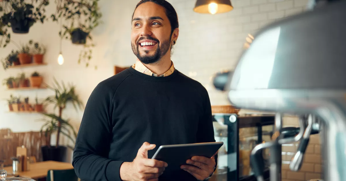 Dono de cafeteria sorrindo com tablet na mão ao lado da máquina de espresso, com plantas e prateleiras de madeira ao fundo