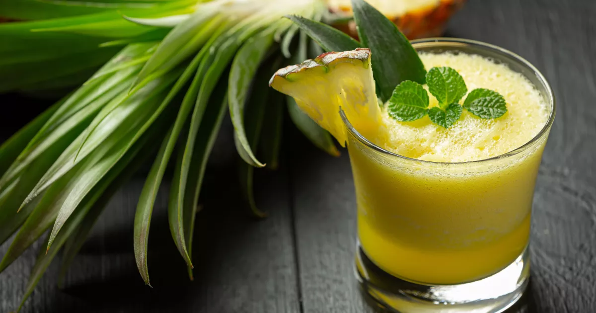 Copo baixo com drink amarelo de abacaxi e espuma, decorado com fatia de abacaxi e hortelã, ao lado de abacaxi inteiro sobre superfície de madeira escura