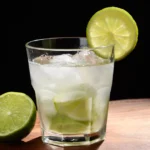 Copo old fashioned com caipirinha sem álcool, pedaços de limão, gelo e rodela de limão na borda, ao lado de meio limão sobre tábua de madeira