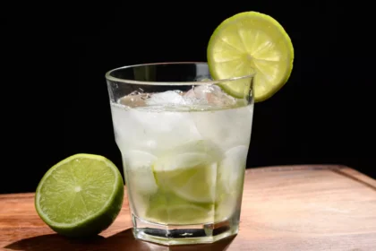 Copo old fashioned com caipirinha sem álcool, pedaços de limão, gelo e rodela de limão na borda, ao lado de meio limão sobre tábua de madeira