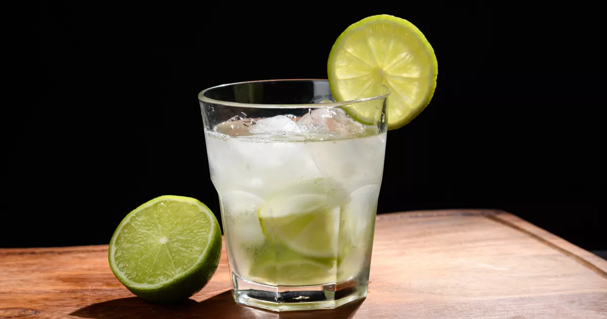 Copo old fashioned com caipirinha sem álcool, pedaços de limão, gelo e rodela de limão na borda, ao lado de meio limão sobre tábua de madeira