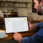 Homem de camisa jeans segurando tablet com o sistema ControleNaMão aberto na tela de comandas, apoiado no balcão de uma cafeteria com croissants e café ao lado e outro funcionário ao fundo