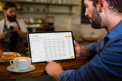 Homem de camisa jeans segurando tablet com o sistema ControleNaMão aberto na tela de comandas, apoiado no balcão de uma cafeteria com croissants e café ao lado e outro funcionário ao fundo