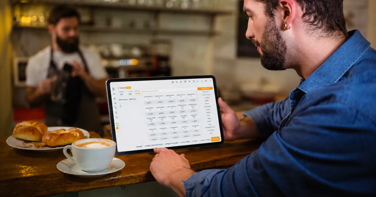 Homem de camisa jeans segurando tablet com o sistema ControleNaMão aberto na tela de comandas, apoiado no balcão de uma cafeteria com croissants e café ao lado e outro funcionário ao fundo
