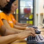 Dois profissionais de camiseta laranja com o logo do CNM trabalham lado a lado em computadores de mesa durante a implantação do sistema para restaurante