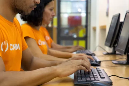 Dois profissionais de camiseta laranja com o logo do CNM trabalham lado a lado em computadores de mesa durante a implantação do sistema para restaurante