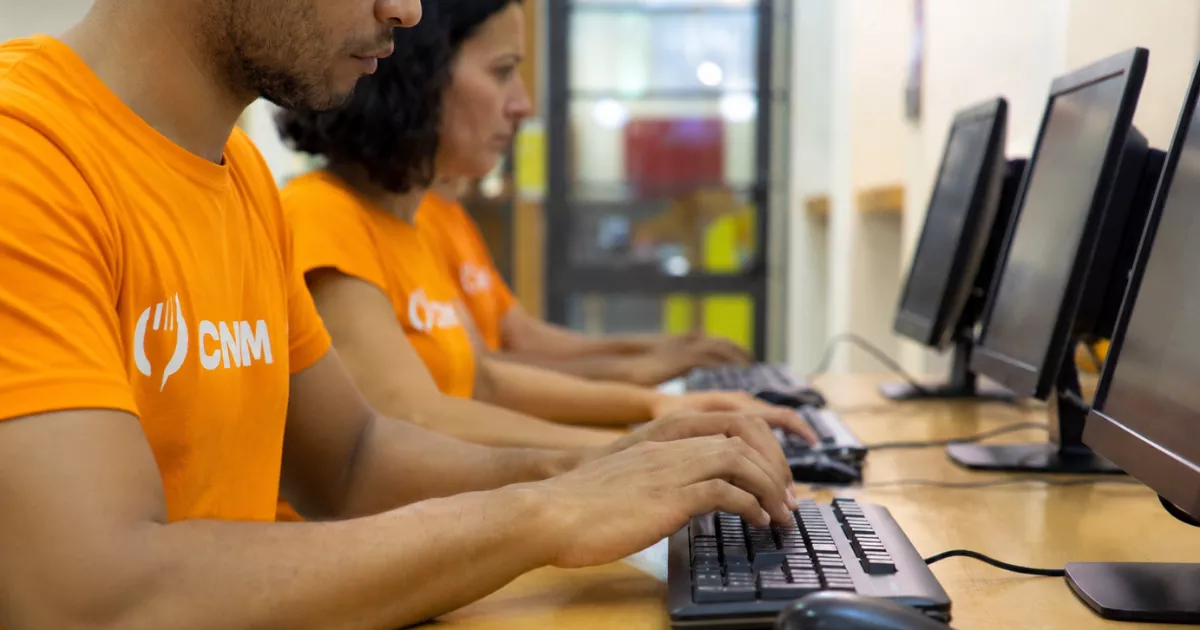 Dois profissionais de camiseta laranja com o logo do CNM trabalham lado a lado em computadores de mesa durante a implantação do sistema para restaurante