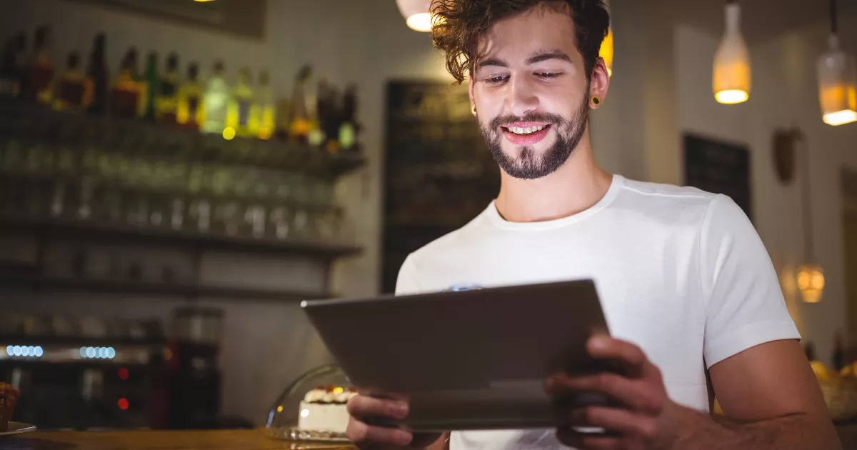 Jovem com barba e camiseta branca sorri enquanto consulta um tablet no balcão de um bar com prateleiras de bebidas e luminária quente ao fundo, monitorando os custos da operação.