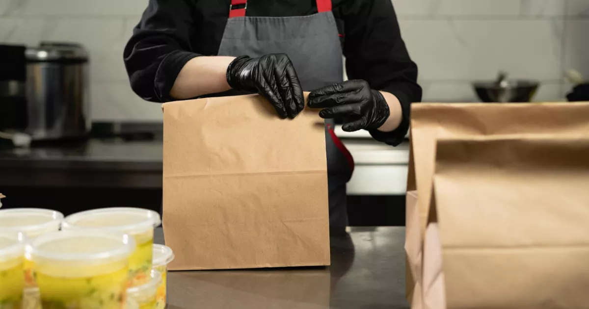 Cozinheiro com avental cinza e luvas pretas fechando sacola kraft sobre bancada de inox, com potes plásticos transparentes de alimento ao lado, na expedição de pedidos de delivery