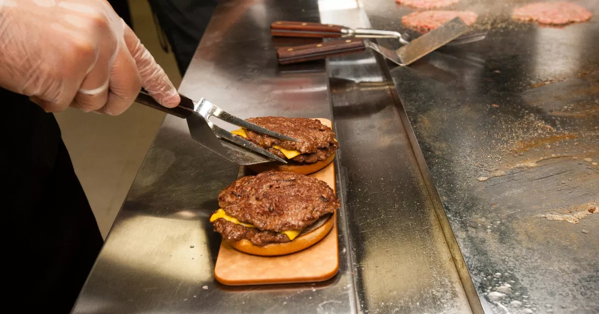 Mãos com luvas manuseando pinça e espátula sobre chapa de inox com hambúrgueres grelhados e queijo derretido, em cozinha de hamburgueria delivery