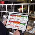 Cozinheiro de uniforme escuro segurando tablet com sistema KDS exibindo fila de pedidos coloridos, em cozinha profissional com bancadas de inox ao fundo