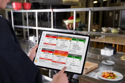 Cozinheiro de uniforme escuro segurando tablet com sistema KDS exibindo fila de pedidos coloridos, em cozinha profissional com bancadas de inox ao fundo
