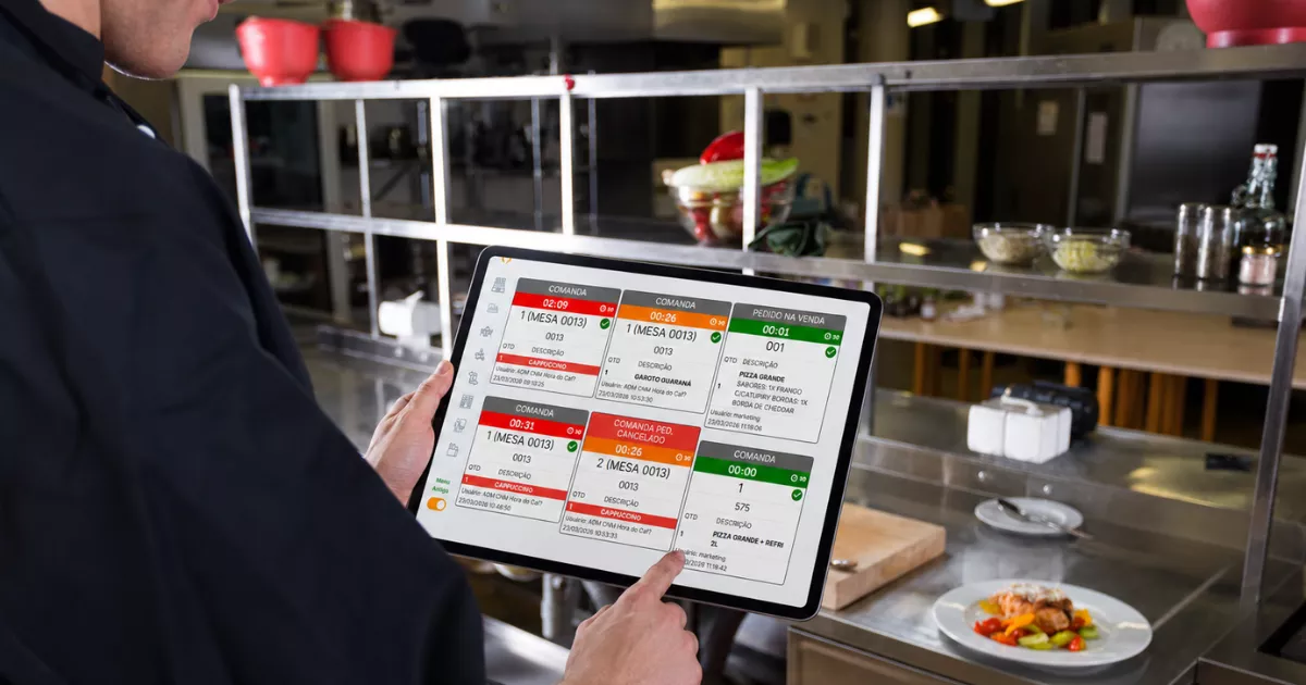 Cozinheiro de uniforme escuro segurando tablet com sistema KDS exibindo fila de pedidos coloridos, em cozinha profissional com bancadas de inox ao fundo