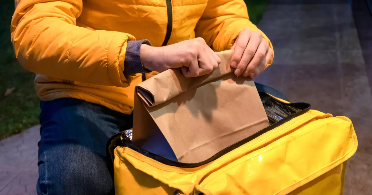 Entregador com jaqueta amarela acomodando sacola kraft dentro de bag térmica amarela aberta, ao ar livre à noite, durante entrega de delivery de restaurante