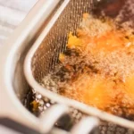 Cesto aramado de inox com alimento empanado dourando em óleo borbulhante dentro da cuba de uma fritadeira industrial, evidenciando o processo de fritura em alta temperatura.