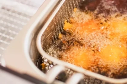 Cesto aramado de inox com alimento empanado dourando em óleo borbulhante dentro da cuba de uma fritadeira industrial, evidenciando o processo de fritura em alta temperatura.
