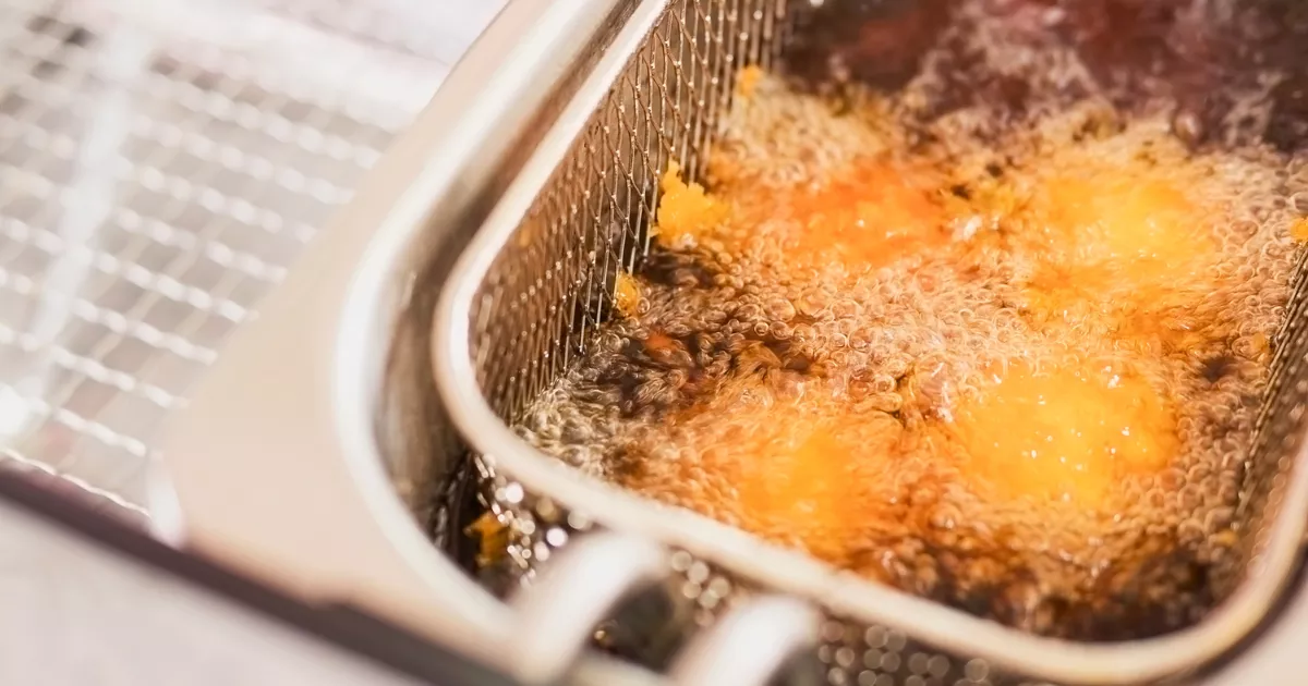 Cesto aramado de inox com alimento empanado dourando em óleo borbulhante dentro da cuba de uma fritadeira industrial, evidenciando o processo de fritura em alta temperatura.