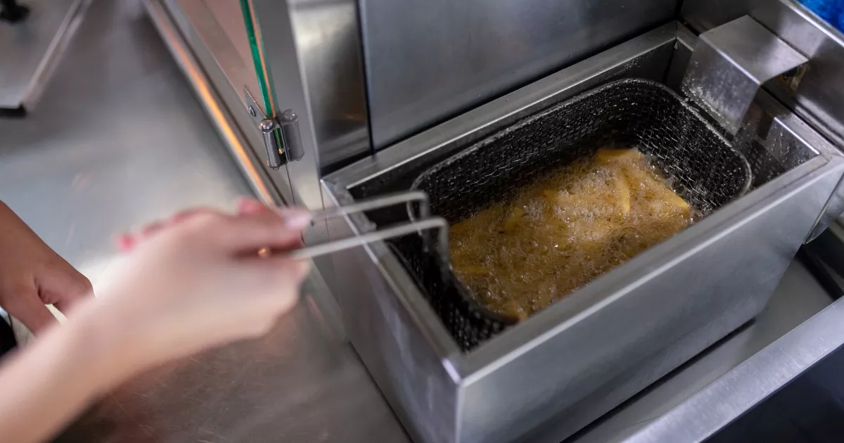 Mãos segurando uma pinça para ajustar o cesto com alimento frito dentro de uma fritadeira elétrica industrial em inox, vista de cima com a tampa aberta em cozinha profissional.