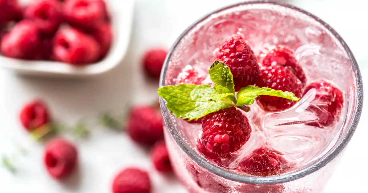 Vista de cima de copo com drink rosado, framboesas frescas flutuando entre cubos de gelo e folha de hortelã, com mais framboesas espalhadas ao redor