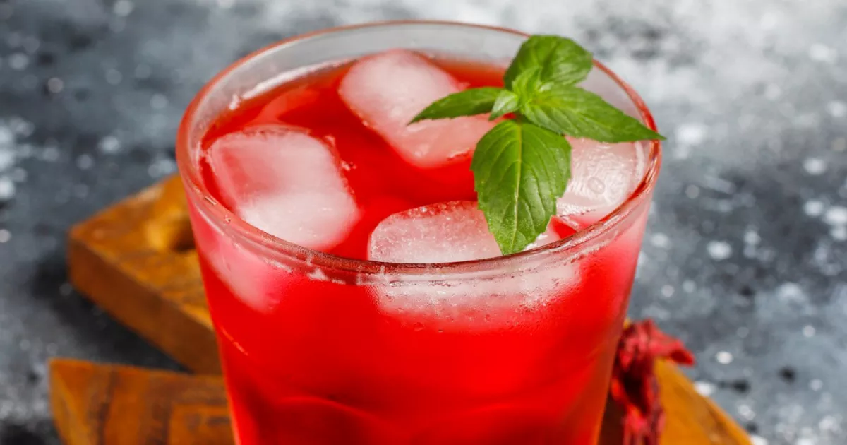 Copo de vidro com drink vermelho intenso de hibisco, cubos de gelo e ramo de hortelã fresca, sobre tábua de madeira com flores secas de hibisco ao lado