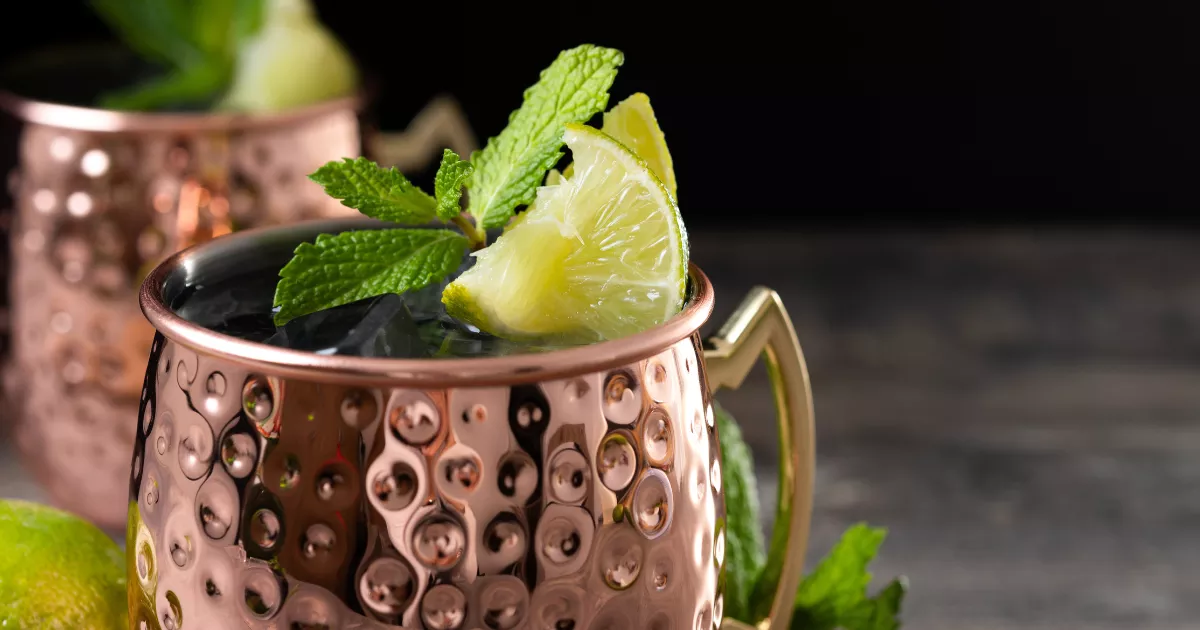 Caneca de cobre martelada com drink decorado com ramo de hortelã e fatia de limão, ao lado de limões frescos sobre superfície escura