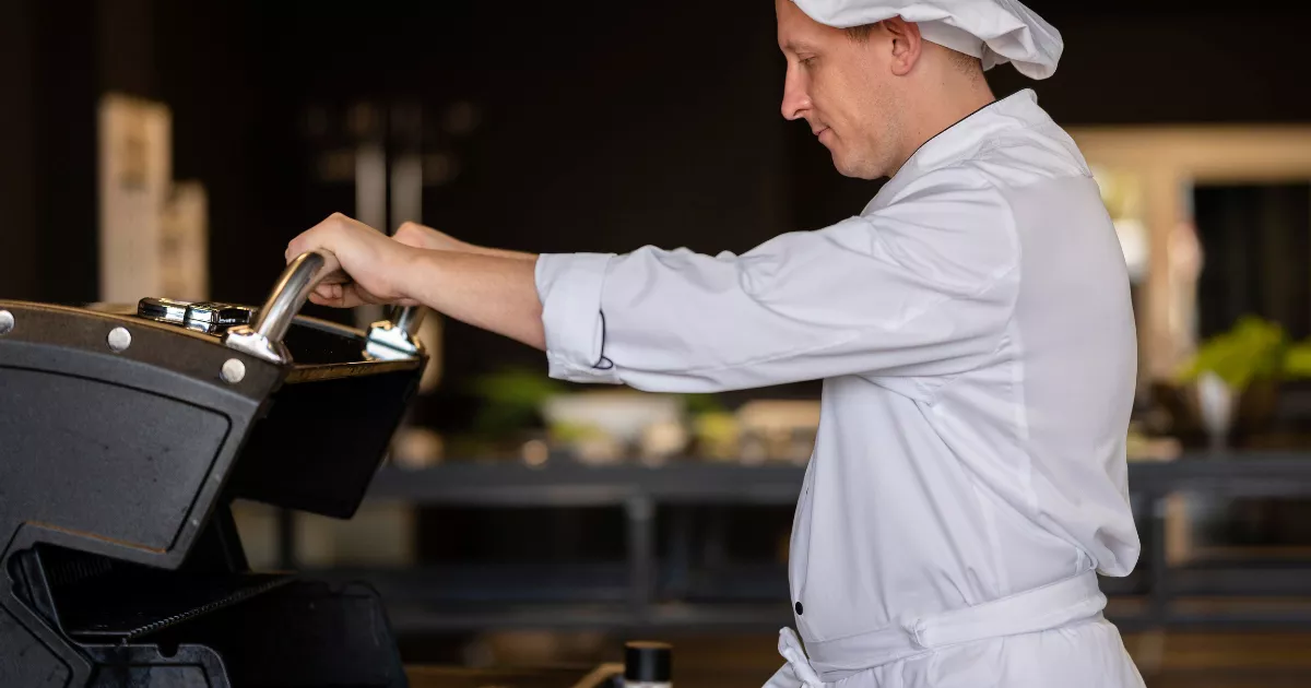 Chef de uniforme e chapéu brancos de perfil, erguendo com as duas mãos a tampa de um equipamento de cocção comercial preto em cozinha profissional, comparando custos de operação.