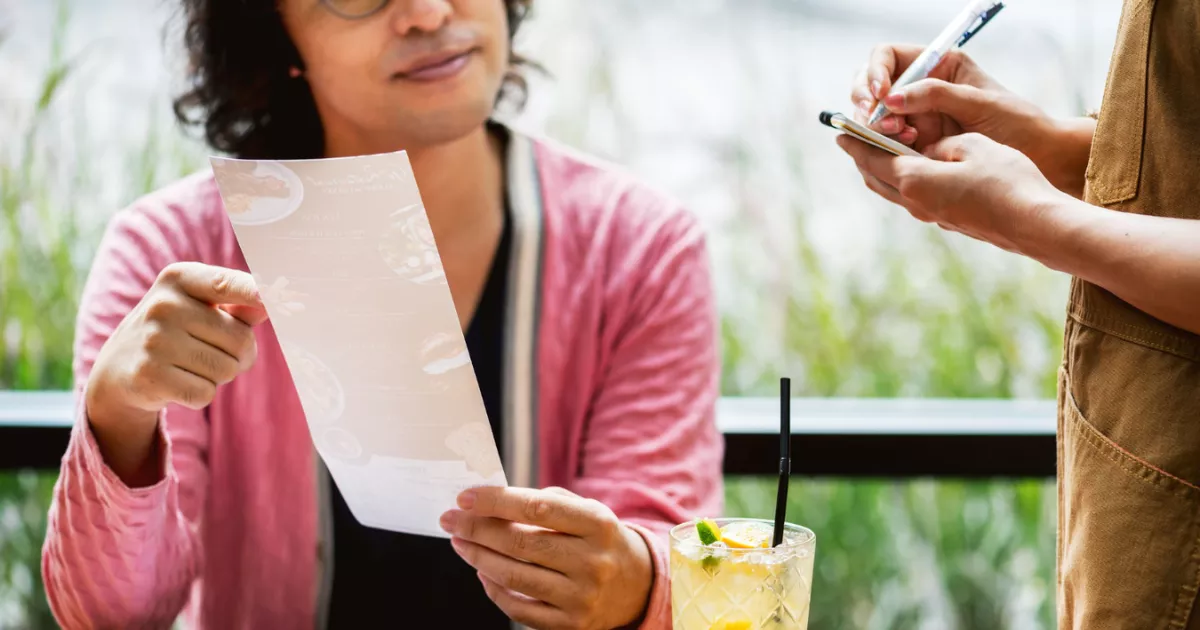 Cliente de óculos segurando cardápio impresso enquanto garçom de avental anota o pedido em bloco de papel, com copo de drink de limão sobre a mesa em restaurante com vegetação ao fundo