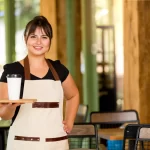 Atendente sorridente com avental claro segurando copo de café em bandeja de madeira, em ambiente acolhedor com móveis rústicos — ideal para ilustrar nomes de cafeterias regionais ou com proposta afetiva.