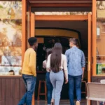 Quatro jovens entrando em restaurante de fachada de madeira e paredes de tijolo, com mesas externas ocupadas à esquerda, ilustrando o movimento de clientes num ponto comercial.
