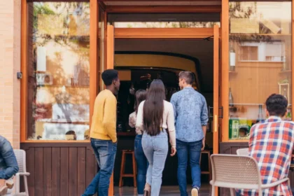 Quatro jovens entrando em restaurante de fachada de madeira e paredes de tijolo, com mesas externas ocupadas à esquerda, ilustrando o movimento de clientes num ponto comercial.