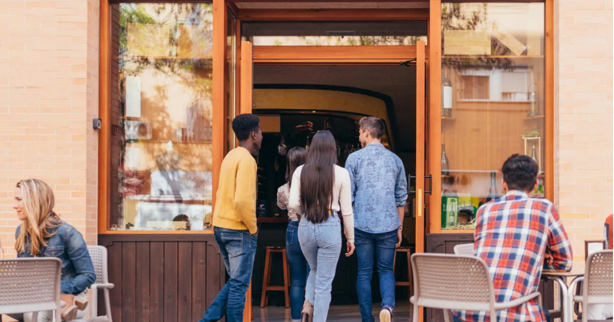 Quatro jovens entrando em restaurante de fachada de madeira e paredes de tijolo, com mesas externas ocupadas à esquerda, ilustrando o movimento de clientes num ponto comercial.