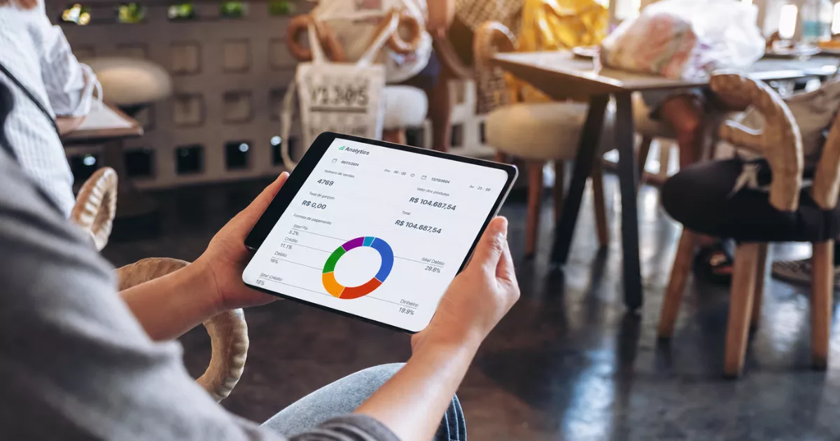 Mãos segurando tablet com painel de analytics que exibe 4.769 vendas e faturamento de R$ 104.687,54, com mesas e clientes de um restaurante visíveis ao fundo.
