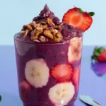 Copo transparente cheio de açaí cremoso com fatias de banana e morangos visíveis pelas laterais, granola crocante por cima e morango fresco na borda, sobre fundo azul claro