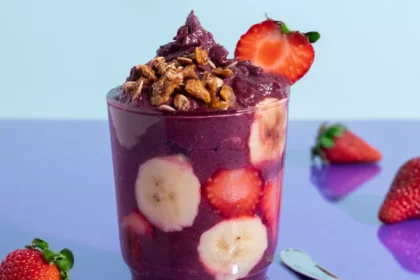 Copo transparente cheio de açaí cremoso com fatias de banana e morangos visíveis pelas laterais, granola crocante por cima e morango fresco na borda, sobre fundo azul claro