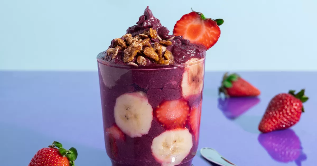 Copo transparente cheio de açaí cremoso com fatias de banana e morangos visíveis pelas laterais, granola crocante por cima e morango fresco na borda, sobre fundo azul claro
