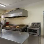 Cozinha de lanchonete de bairro com chapa, fritadeira dupla, coifa de inox e geladeira expositora abastecida, mostrando os principais equipamentos em operação real