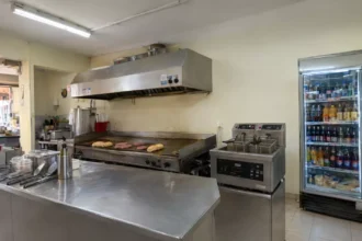 Cozinha de lanchonete de bairro com chapa, fritadeira dupla, coifa de inox e geladeira expositora abastecida, mostrando os principais equipamentos em operação real