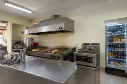 Cozinha de lanchonete de bairro com chapa, fritadeira dupla, coifa de inox e geladeira expositora abastecida, mostrando os principais equipamentos em operação real