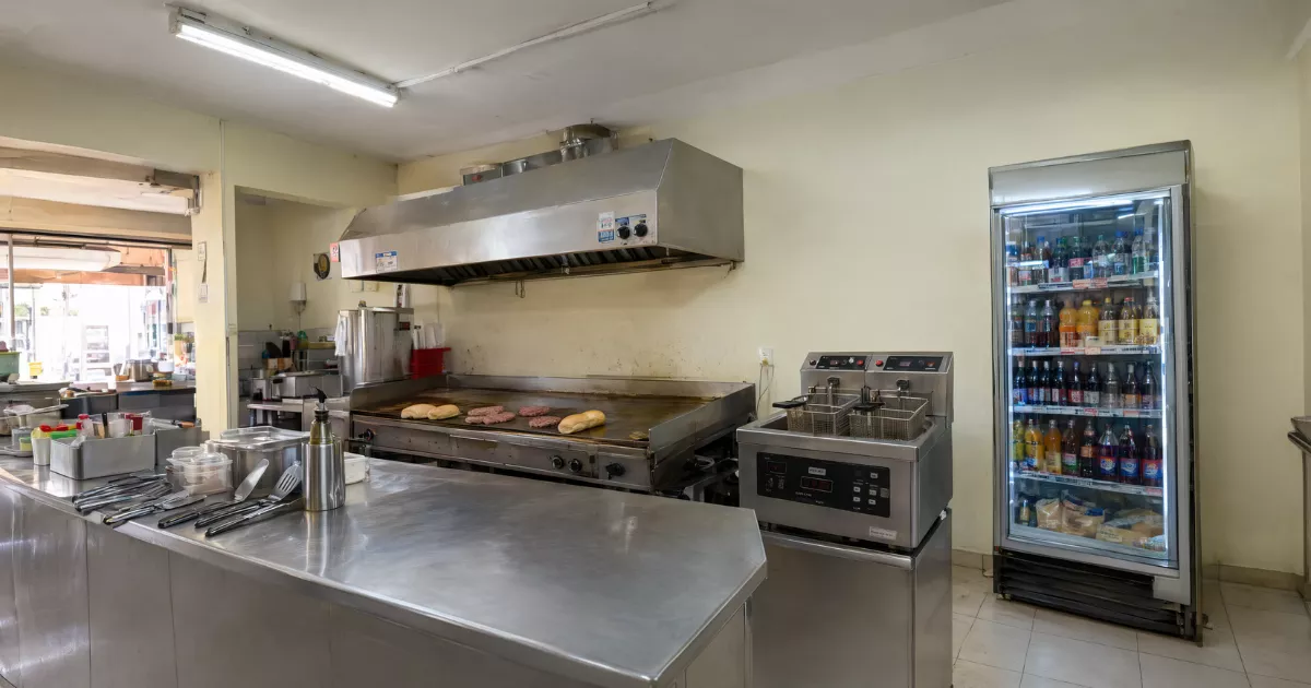 Cozinha de lanchonete de bairro com chapa, fritadeira dupla, coifa de inox e geladeira expositora abastecida, mostrando os principais equipamentos em operação real