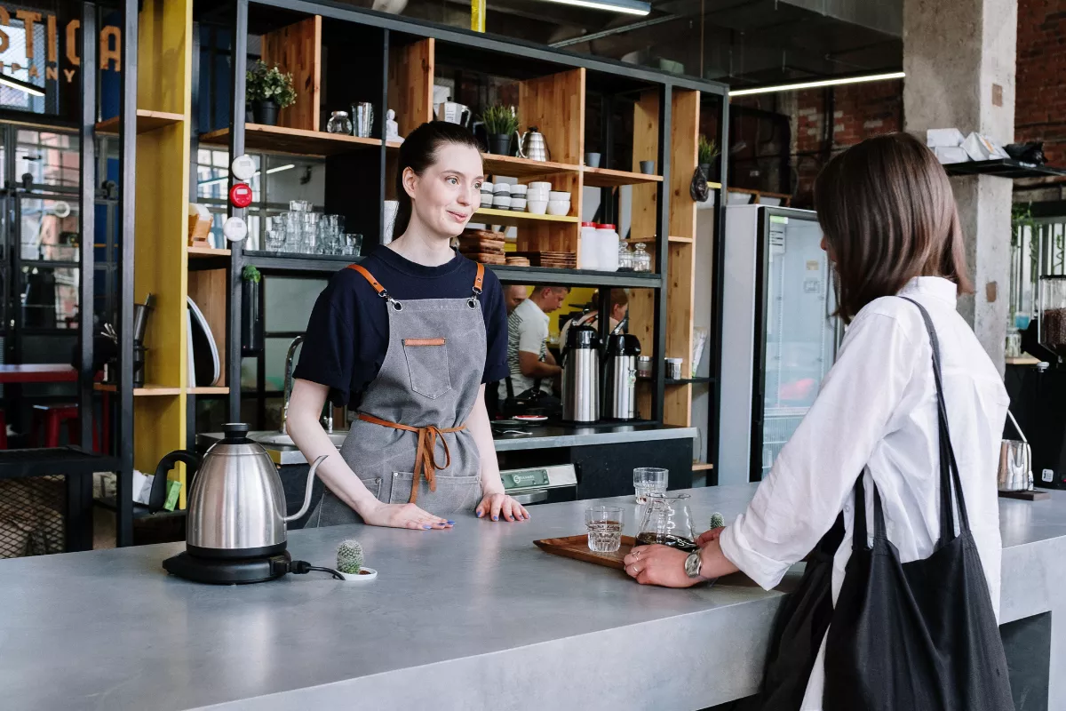 Barista com avental atendendo cliente em cafeteria moderna, com prateleiras organizadas ao fundo e utensílios de preparo à vista — representando atendimento personalizado e conceito de marca para cafeterias urbanas.